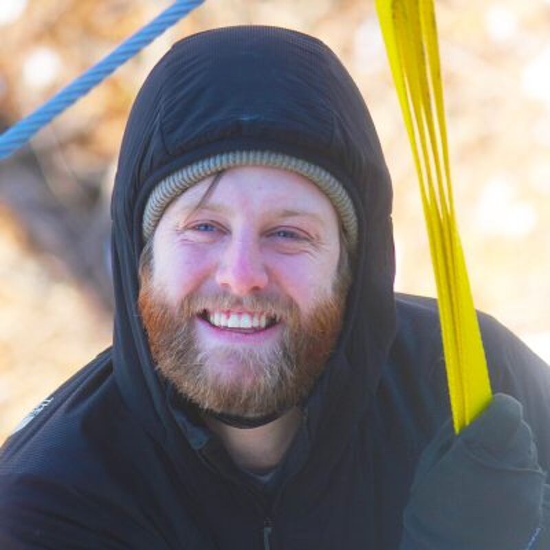 The image shows a man with a red beard and a beanie under a black hood. He is smiling and holding a yellow object. The background appears to be outdoors, possibly in a snowy environment. The man is wearing black gloves. The overall impression is of a person enjoying an outdoor activity.